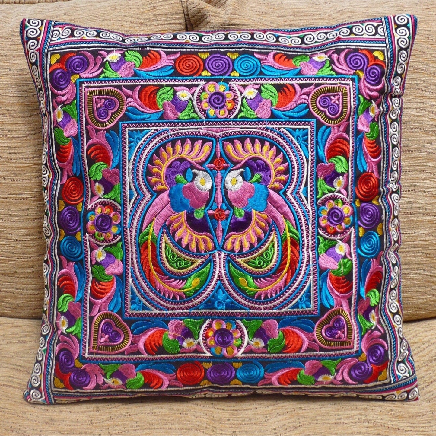 A brightly colored, embroidered cushion cover featuring geometric patterns and a central face motif, displayed on a neutral background.