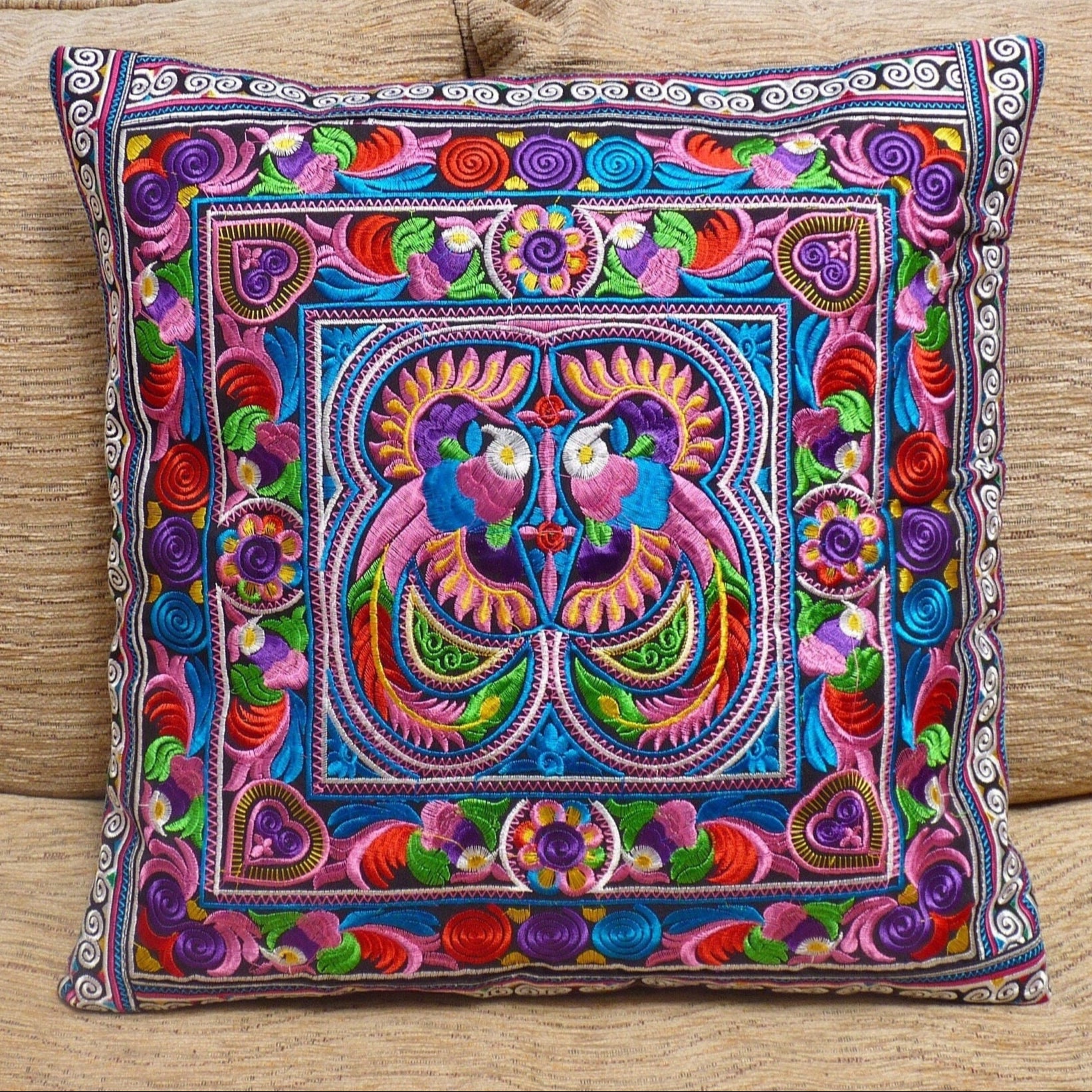 A brightly colored, embroidered cushion cover featuring geometric patterns and a central face motif, displayed on a neutral background.