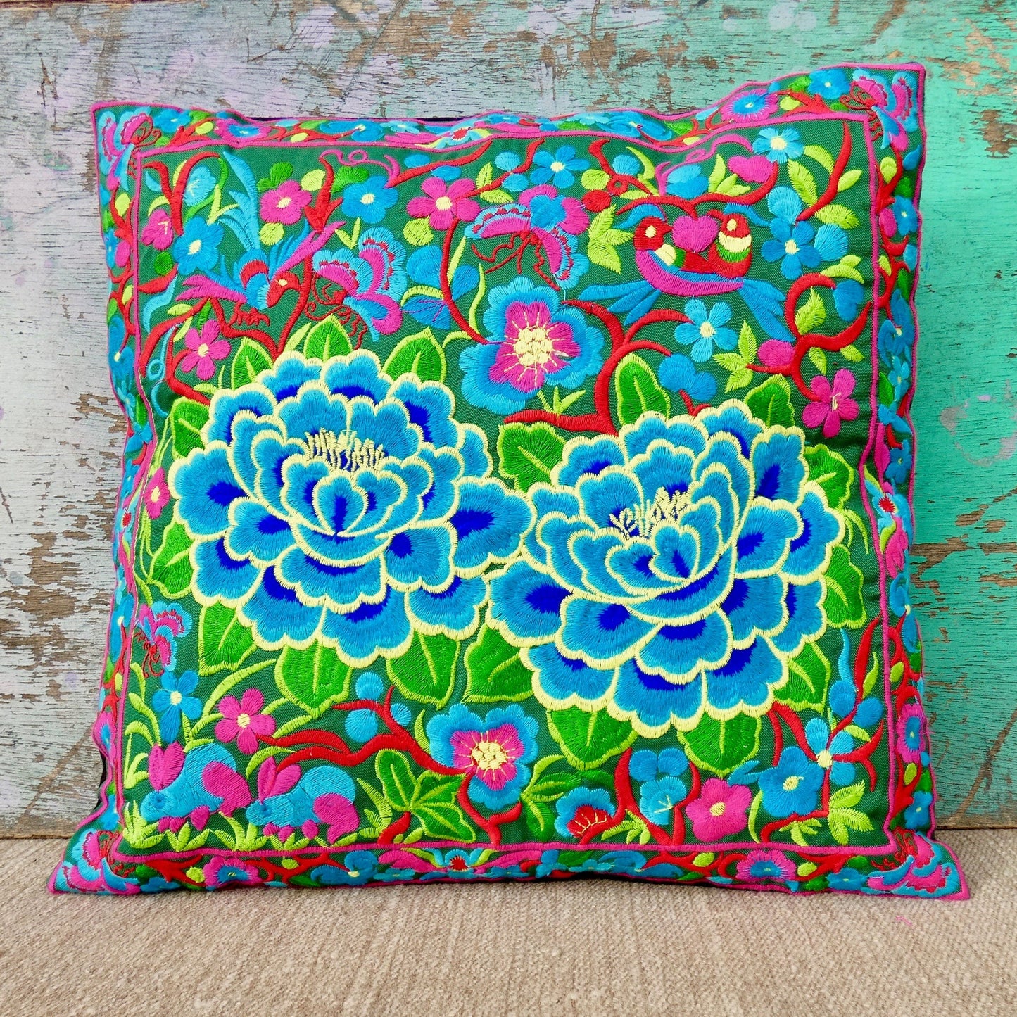 A colorful cushion cover with a vibrant floral embroidery design, featuring blue, pink, and green colors on a wooden surface.