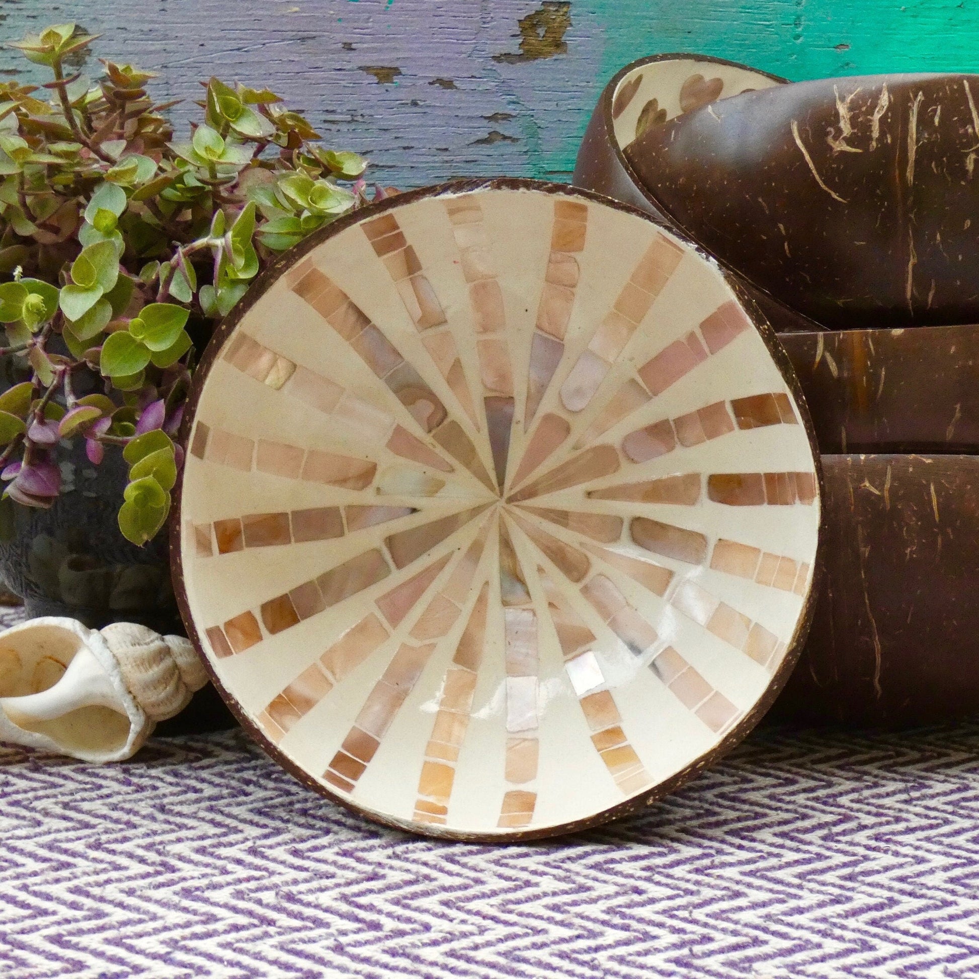 A coconut bowl with mother of pearl inlay, placed on a patterned surface, with a plant and a coconut shell in the background.
