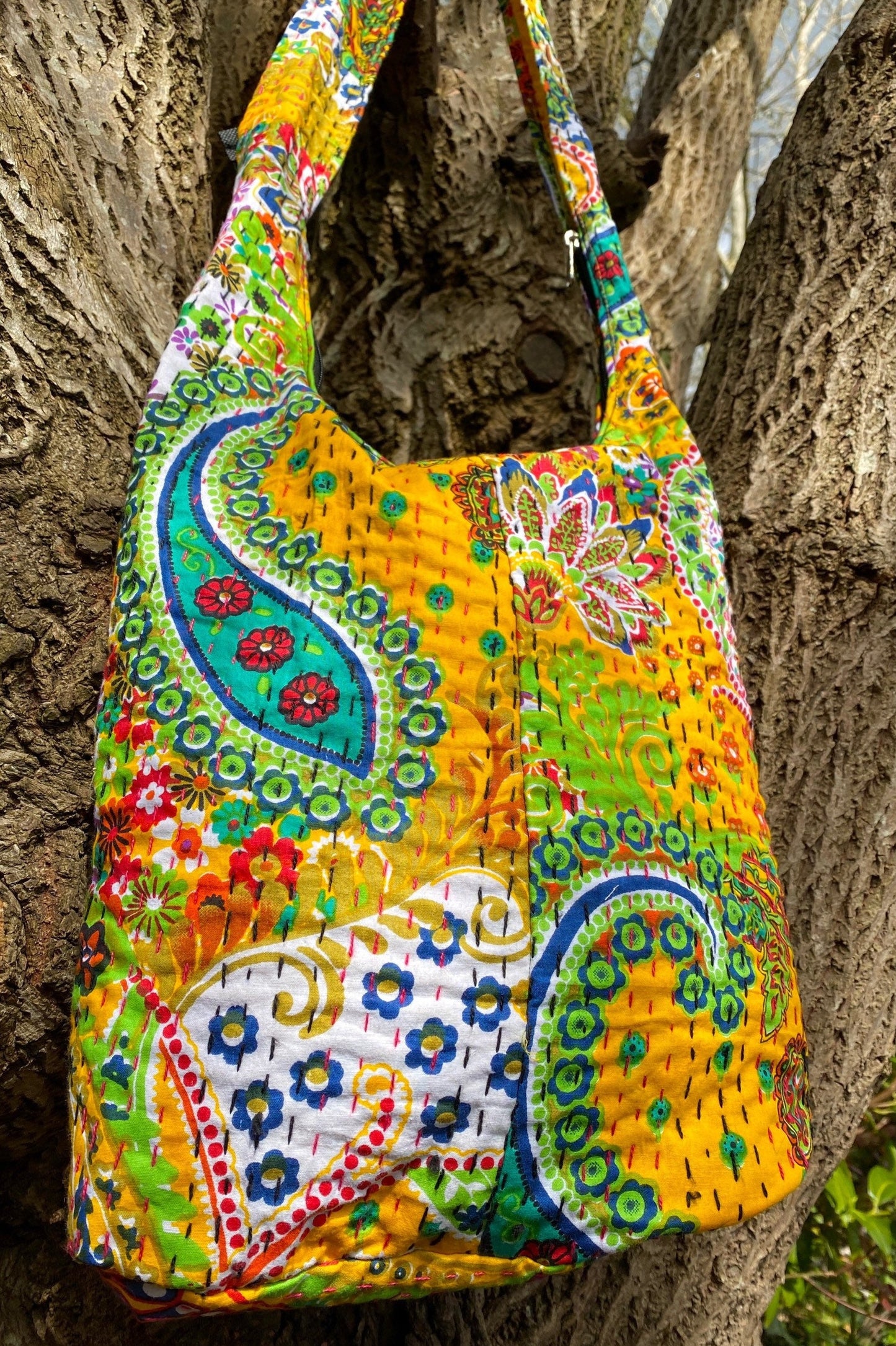 A colorful handmade Indian Kantha fabric bucket bag displayed hanging on a tree.