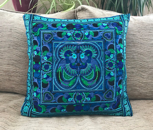 A vibrant turquoise cushion cover with a bird of paradise design, featuring traditional Hmong embroidery, displayed on a neutral sofa background.